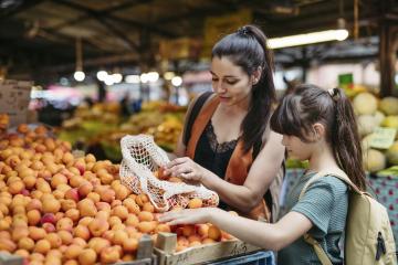 A woman and adolescent girl, both with long brown ponytails, load up a netted market bag with apricots at an open-air farmer's market structure.