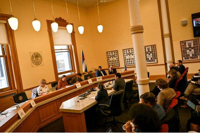 People seated around a curved committee dais during a legislative hearing in a large government meeting room with high ceilings, tall windows, and a Colorado state flag.