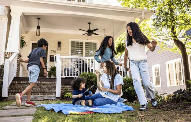 A family plays together in the front yard of a suburban home. An adult woman sits on a blanket blowing bubbles for a toddler, while two older children and another woman laugh and move around them near the porch of the house.