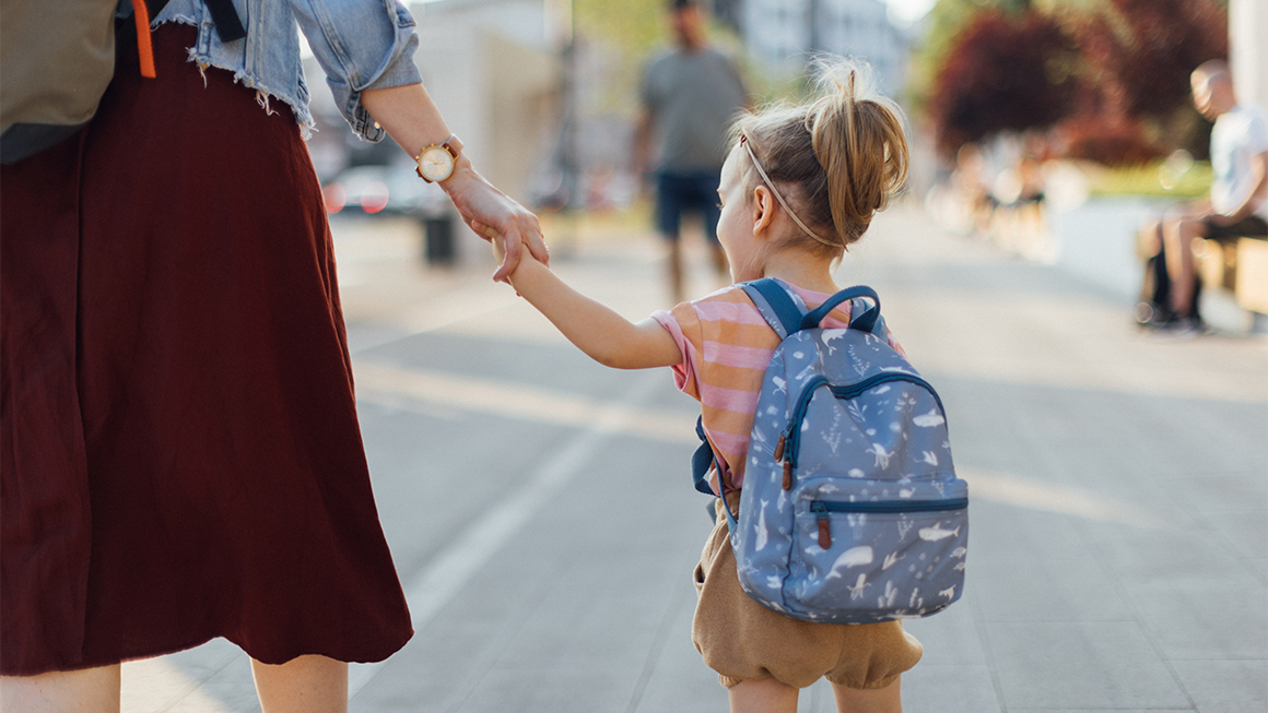 A young child wearing a small blue backpack holds an adult’s hand while walking down a sunlit city sidewalk