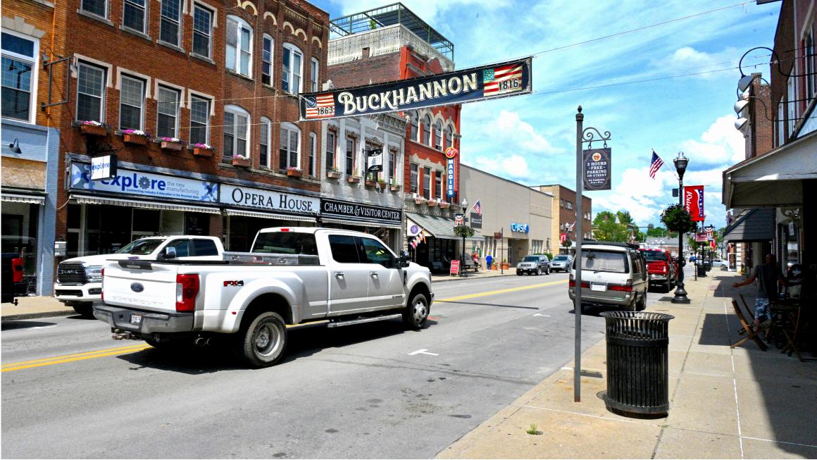 Downtown street in Buckhannon, West Virginia, with brick storefronts, parked vehicles, and a large “Buckhannon” banner hanging above the road.
