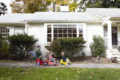 A dad and his two sons riding toy cars in front of their home. 