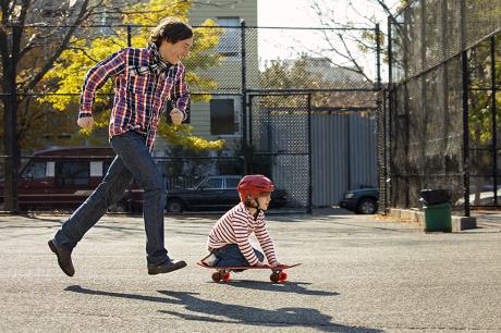 A dad playing outside with his child while they ride a skateboard.