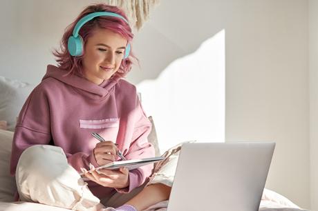 A young girl with pink hair and headphones working on a laptop. 