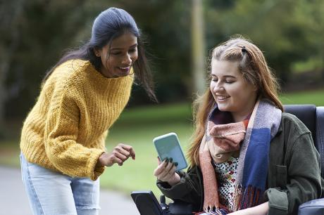 A young woman in a wheelchair talking to another woman and looking at phone together.
