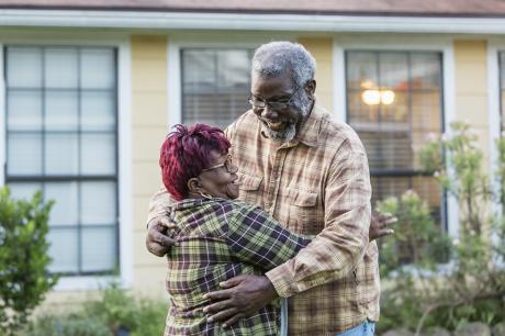 An older black couple hugging in front of housing.