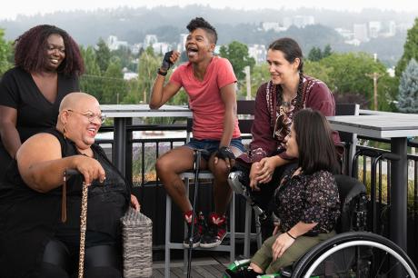 Five disabled people of color with canes, prosthetic legs, and a wheelchair sit on a rooftop deck, laughing and sharing stories. Greenery and city high-rises are visible in the background.