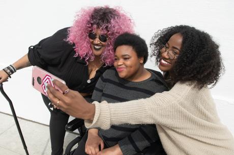 hree Black and disabled friends (a non-binary person with a cane and tangle stim toy, a non-binary person in a power wheelchair, and an invisibly disabled femme) smiling and taking a cell phone selfie together. The photo is angled from the left and cropped around everyone's torso.