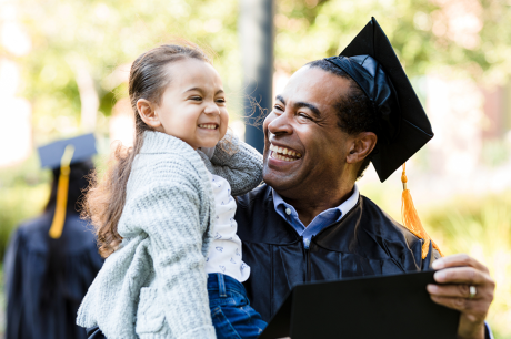 A dad graduating with a cap and gown holding his daughter outside.