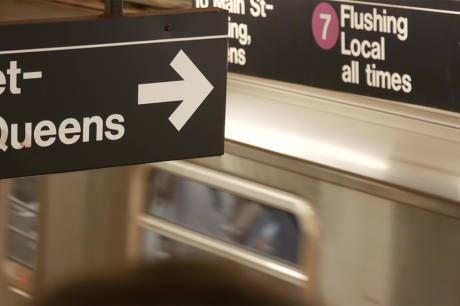 Photo of the inside of a New York subway station, as a subway car is going by. A black sign in the foreground reads "Queens" and points to the platform. Another sign in the distance reads "7: Flushing, Local all times."