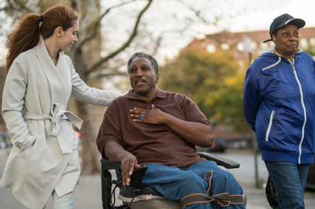 A photo of a young white woman, an older Black man in a wheelchair, and an older Black woman talking as they stroll and roll down the sidewalk together.