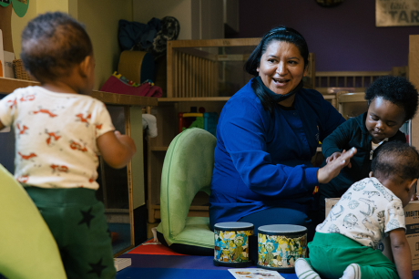 Photo of a child care worker sitting on the ground and smiling as she plays with three toddlers. 