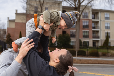 photo of a woman laughing while playfully lifting her one year old toddler son up in the air as her husband affectionately watches