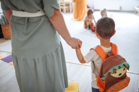 Photo of a toddler wearing a backpack holding his mom's hand as they arrive at daycare.