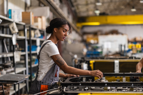 Photo of a young Black woman working on an electric car engine in a factory.