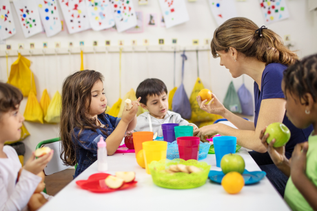 Photo of a young white woman sitting at a table with a diverse group of preschoolers as they eat fruit. 
