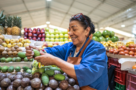 Photo of an older woman restocking avocados at a grocery store.