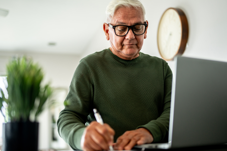 Image of an older man in a green shirt writing something down on a paper next to his laptop.