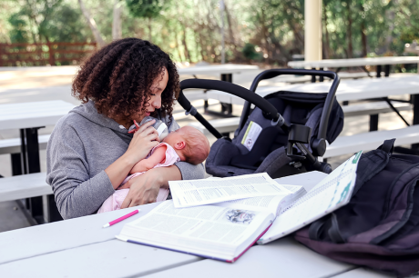 A young mom studies for a college exam outside at a picnic table while feeding her baby a bottle
