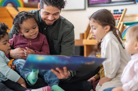 A daycare educator sits on the floor with a child on his lap and other toddlers scattered around him as he reads them a story. They are each dressed casually and are focused on the book.
