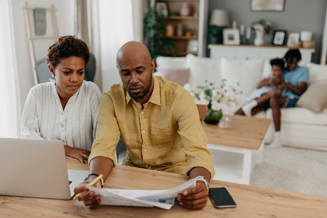 A husband and wife sit at a table and review paperwork together. Their kids sit on a couch playing behind them.