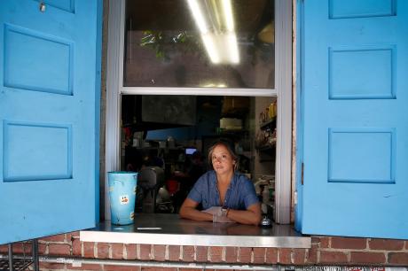 A woman working in a restaurant looks out a carry-out window at the camera