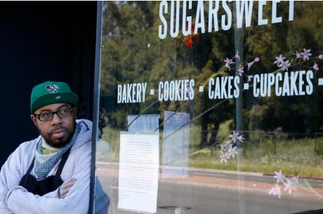 A man and small business owner leans against the doorway of a bakery looking off into the distance