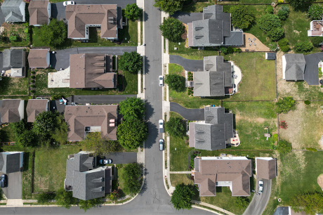 Aerial top-down view of a suburban neighborhood with single-family houses, green lawns, driveways, and trees lining a grid of residential streets, with a few cars parked along the road.