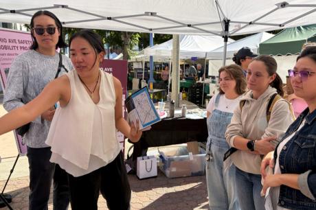 A woman at an outdoor community event stands under a white canopy tent, pointing to a display board while holding a clipboard with a map. Several people stand around her listening. Behind them are tables with materials and a pink sign that reads “Ohio Ave Safety Mobility Project.”