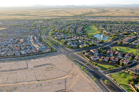 Aerial view of a large suburban neighborhood in a desert landscape, with curving residential streets, single-family homes, a small lake with a fountain, and a golf course, bordered by open undeveloped land and distant mountains.