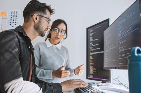 Photo of two young professionals looking at lines of code on two monitors.