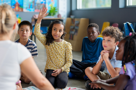 Photo of a young girl of color raising her hand as she and other students sit on a rug with their teacher. 