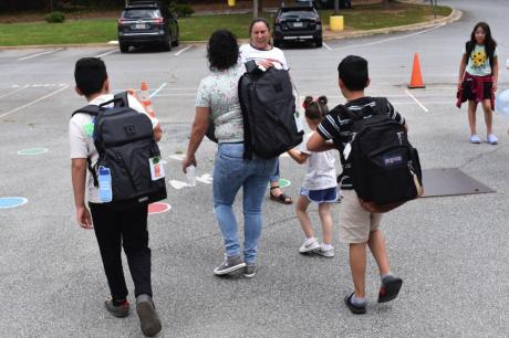 A family of adults and children wearing backpacks walks away in a parking lot 