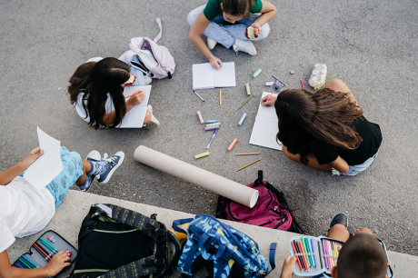 A group of children sit outdoors on pavement in a circle, drawing in notebooks with colorful pencils and chalk scattered around them. Backpacks and art supplies lie nearby, and one child holds a piece of paper while another eats an apple.