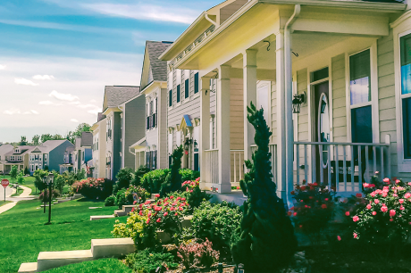 A quiet suburban street with modern two‑story houses, each with front porches, trimmed lawns, and colorful landscaped gardens. 