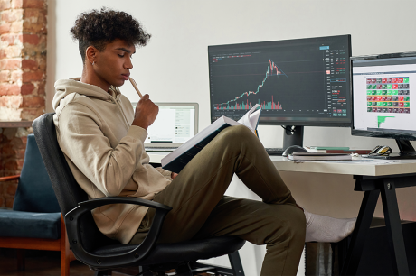 a young man working at a desktop computer at home, reading a book while analyzing financial charts on the screen