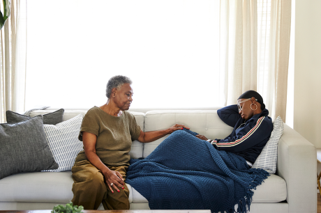 An older adult sits on a couch gently resting a hand on a younger person who is lying under a blue blanket