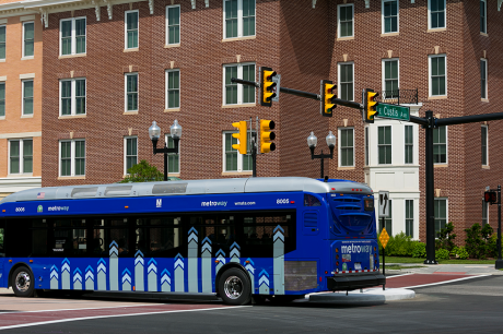 Blue Metroway city bus stopped at an intersection with traffic lights in front of a red brick building.
