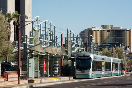 Light rail train arriving at a modern outdoor station with curved canopy structures and city buildings in the background.