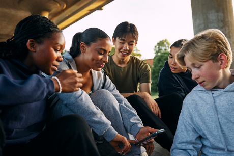 Five young people sit together outside, gathered closely around a smartphone.