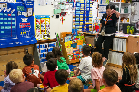 An educator stands at the front of a colorful classroom, reading to a group of young children seated on a carpet, surrounded by learning charts, books, and classroom displays.