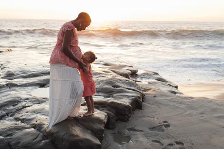 Pregnant person stands with child near ocean