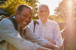 Two older men smile at the camera while leaning against a fence