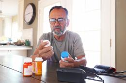 An elderly man in a grey shirt and tortoiseshell glasses sits at home in front of a blood pressure cuff and several prescription bottles, reading labels. 