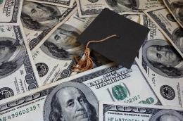 A miniature graduation cap with an orange tassel sits on top of a bunch of American $100 bills.