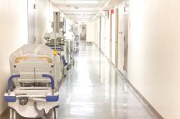 A brightly lit, clean hospital corridor with equipment and portable hospital beds lining the left wall.