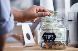 A close-up of a tip jar with a hand-written label with a few dollars' worth of change and two $20 bills, showing a person out of frame inserting a $1 bill. 