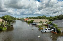 A neighborhood street of single-story homes is deeply flooded though the sky is cloudy and sunny, with water levels covering car tires and mailbox posts.