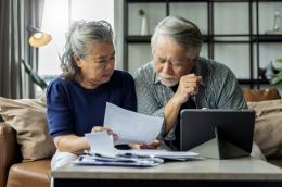 n elderly woman and man sit side-by-side with furrowed brows and looks of worry in front of a tablet and a pile of papers, inspecting an invoice.