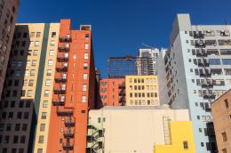 Several multistory, colorful buildings, some with fire escapes, clustered close together are illuminated by the sun.